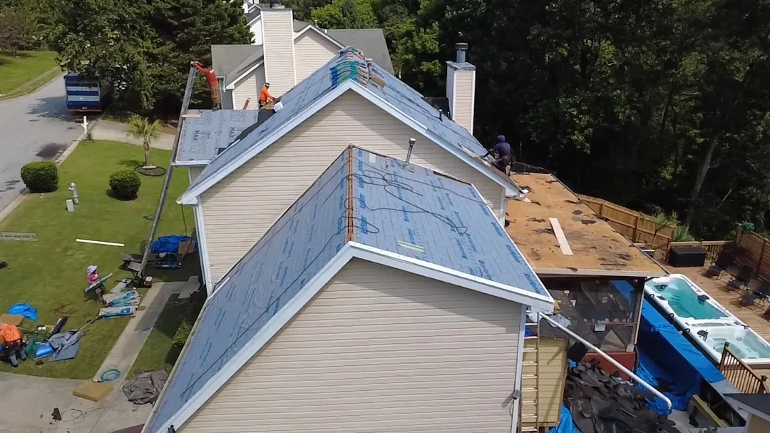 The same home mid-job: synthetic underlayment on the upper slopes, bare decking on the rear section, crew on the roof, and blue tarps over the patio and lawn below.