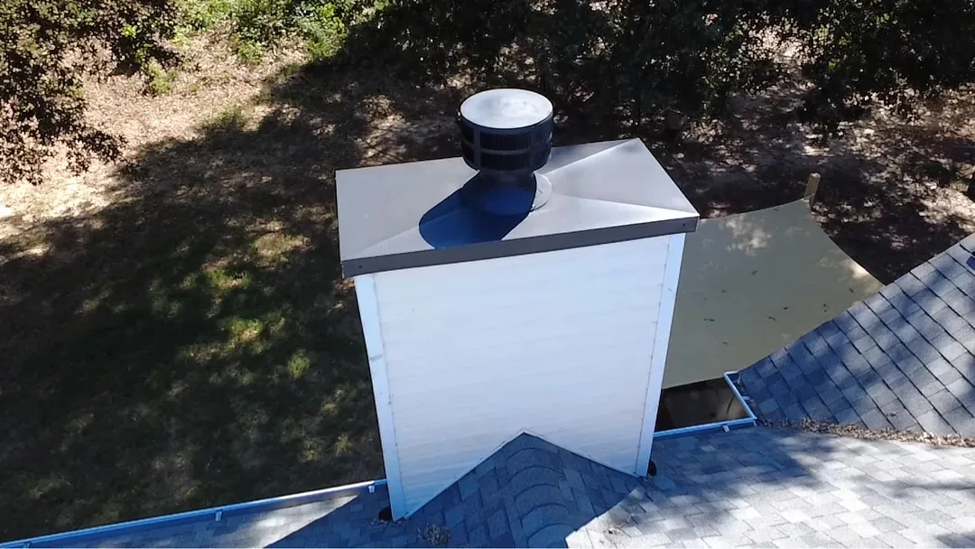 The same chimney with a new metal pan and flue cap fitted, and new gray shingles around its base.