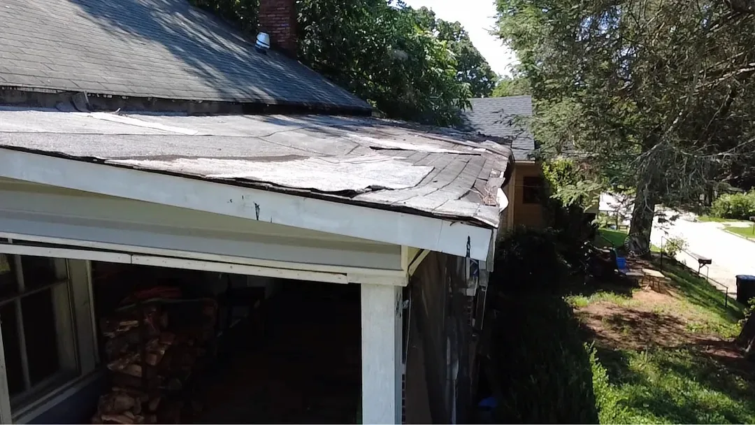 Close view of a porch roof before the work: old roofing lifting and split along the eave above a sagging fascia board.