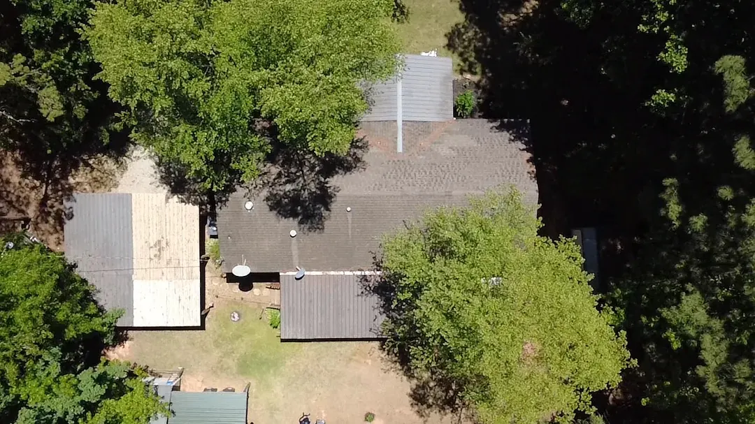 Top-down aerial of a home under heavy tree cover before replacement, its old shingle roof weathered and stained.
