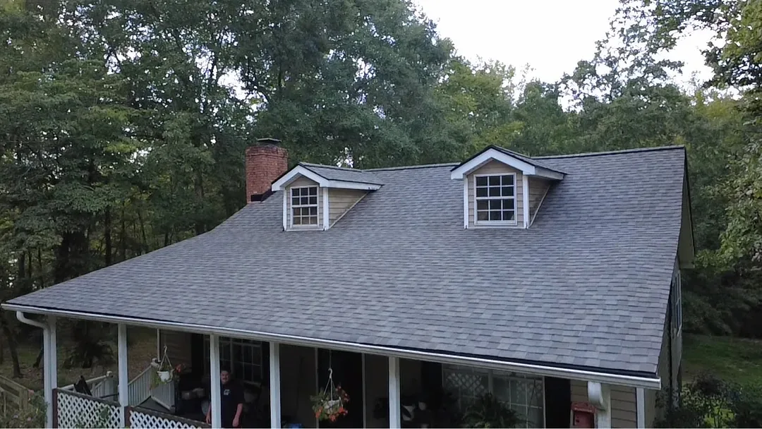 Finished architectural shingle roof in a brown-gray blend on a home with two dormers, a brick chimney and a full-width front porch.
